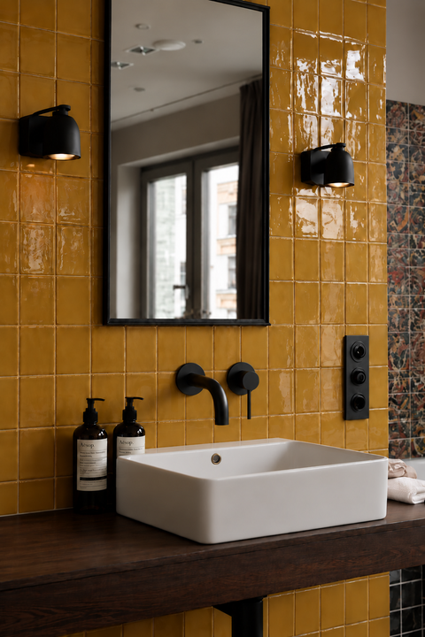 Mitigeur lavabo encastré mural Ysel noir mat Azéo — au-dessus d'une vasque rectangulaire blanche, carrelage jaune moutarde brillant, plan en bois foncé et miroir noir