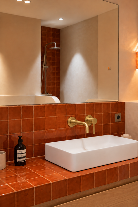 Mitigeur lavabo encastré mural Ysel or brossé Azéo — au-dessus d'une vasque rectangulaire blanche, carrelage terracotta brillant et grand miroir