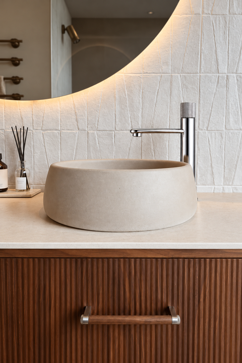 Mitigeur lavabo haut bec plat Ostenne chromé Azéo — à côté d'une vasque ronde en béton clair, meuble cannelé en noyer et miroir rond rétroéclairé