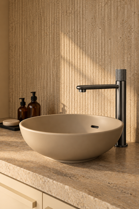 Mitigeur lavabo haut bec plat Ostenne gris anthracite Azéo — à côté d'une vasque ronde beige posée sur un plan en travertin, mur enduit cannelé en lumière rasante