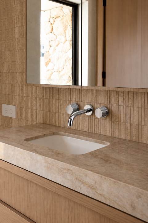 Mitigeur lavabo mural encastré double poignée Havren chromé Azéo — au-dessus d'une vasque encastrée sous un plan en travertin, carrelage beige fin et armoire miroir